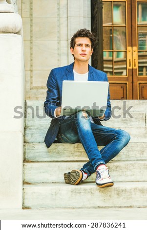 European college student studying in New York. Wearing blue blazer, jeans, sneakers, a young guy sitting on stairs on campus, crossing legs, reading, working on laptop computer. Instagram effect.