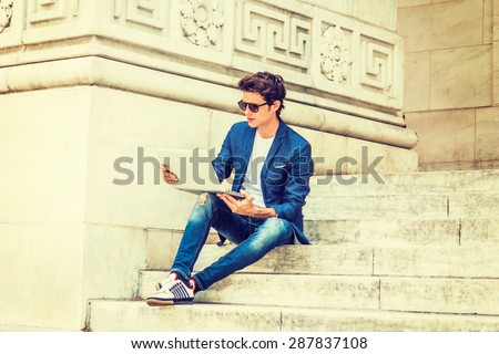 European college student studying in New York. Wearing blue blazer, jeans, sneakers, sunglasses, young guy sitting on stairs on campus, reading, thinking, working on laptop computer. Instagram effect.