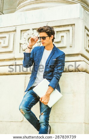European college student studying in New York. Wearing blue blazer, jeans, sunglasses, holding laptop computer, a young guy standing by column on campus, looking down, thinking. Instagram effect.