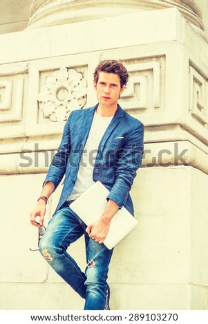 European college student studying in New York. Wearing blue blazer, white under shirt, jeans, holding laptop computer, sunglasses, a young guy standing by column on campus, relaxing. Instagram effect