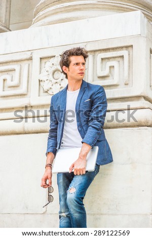 European college student studying in New York. Wearing blue blazer, white under shirt, jeans, holding laptop computer, sunglasses, a young guy standing by column on campus, looking away, relaxing.