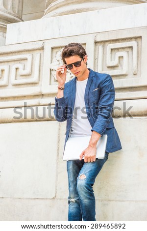European college student studying in New York. Wearing blue blazer, white under shirt, jeans, sunglasses, holding laptop computer, a young guy standing by column on campus, looking down, thinking.