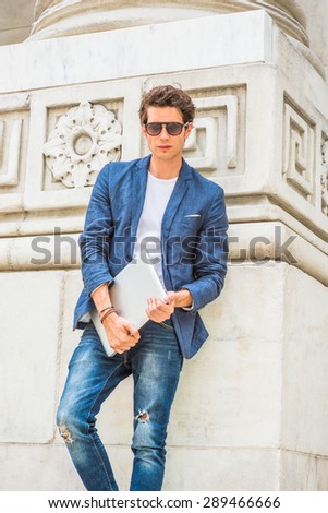 European college student studying in New York. Wearing blue blazer, white under shirt, jeans, sunglasses, holding laptop computer, a young guy standing by column on campus, looking at you, thinking.