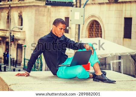 African American businessman working on street in New York. Wearing black shirt, tie, light green pants, leather shoes, young tough black guy reading, working on laptop computer. Instagram effect.
