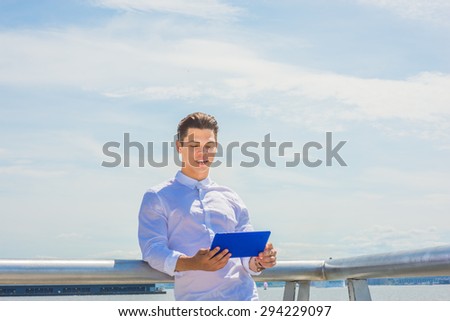 Power of reading. Technology made life easier.  Wearing white shirt, holding blue tablet computer, a college student standing by Hudson River in New York under blue sky, reading, thinking.  Copy Space