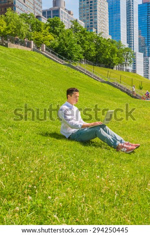 Man traveling, working in New York. Wearing white shirt, jeans, sneakers, a young college student sitting on green lawn with stairs to business district, reading, working on a laptop computer.