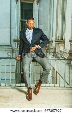 Man waiting for you. Dressing in black blazer, gray pants, brown leather shoes,  African American businessman sitting on railing in vintage style office, looking at his wristwatch. Time is money.