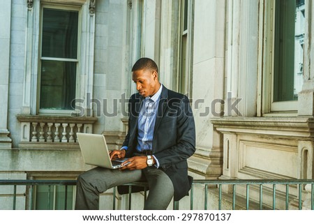 African American businessman working in New York. Wearing black blazer, wristwatch, a guy sitting on railing inside vintage style office building, looking down, reading, working on laptop computer.