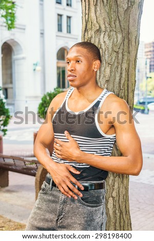 Summer Heat. Man casual fashion. Wearing black, white striped tank top, a young, sexy African American guy leaning against tree trunk, hands resting on stomach, relaxing, thinking, lost in thought.