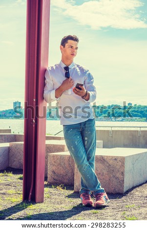 Where are you? Wearing white shirt, jeans, sneakers, sunglasses hanging on collar, young guy standing against pole by Hudson River in New York, holding mobile phone, waiting for you. Instagram effect.