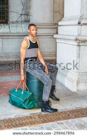 Man casual street fashion. Wearing black, white striped tank top, jeans, leather boots, hand carrying green bag, a young African American guy sitting on street in New York, waiting, relaxing.