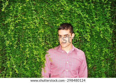 Love Green. Portrait of Student. Wearing red patterned, long sleeve shirt, a young guy standing against wall with long green leaves, smiling, looking at you. Concept of Environment Protection.
