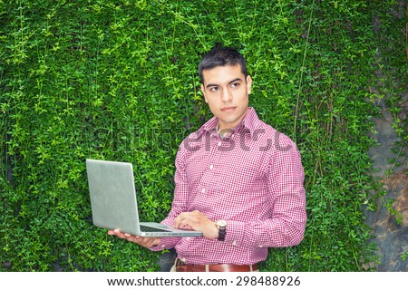 College student studying in peaceful, green environment. Wearing red patterned, long sleeve shirt,  a young guy standing against wall with green leaves on campus, reading, working on laptop computer.