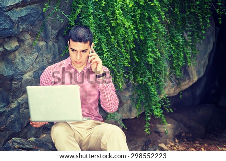 Wearing red, white patterned shirt, a young college student sitting against rocks with green leaves on campus, reading, working on laptop computer, calling on cell phone in same time. Instagram effect