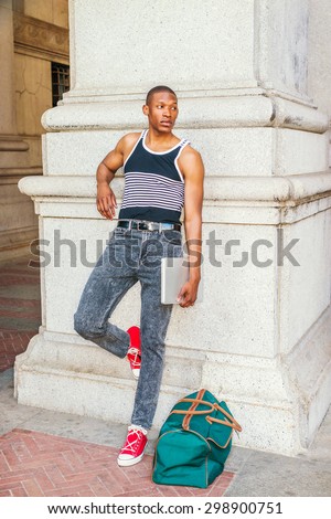 College student traveling in summer in New York. Wearing black, white striped tank top, jeans, red sneakers, carrying green bag, holding laptop computer, a black guy standing against column on street.