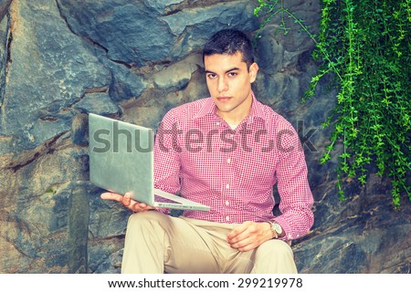 Studying Outside. Wearing red, white patterned shirt, wristwatch, a young college student sitting against rocks with long green leaves on campus, reading, thinking, working on laptop computer.