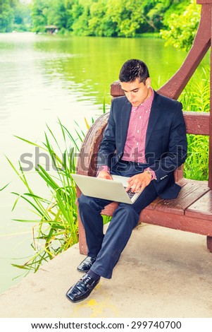 Working hard in peaceful environment. Wearing black suit, red, white patterned undershirt, leather shoes, a young college student sitting by green lake on campus, reading, working on laptop computer.