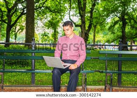 Man working outside. Wearing red, white patterned shirt, black pants, a young college student sitting on chair under trees, reading, working on laptop computer. Technology in our daily life.
