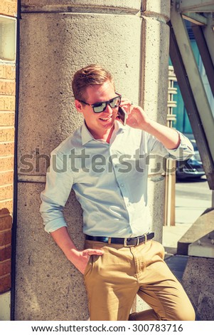 Good to hear from you. Dressing in white shirt, yellow pants, wearing sunglasses, a young guy standing against column on street in New York under strong sunshine, smiling, talking on his mobile phone.