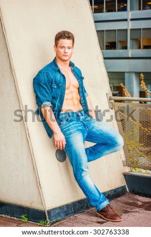 Portrait of hard worker. Wearing Denim shirt, unbuttoned, sleeves rolled, jeans, leather shoes, a young, muscular, sexy guy casually standing against wall on balcony, bending leg, relaxing, thinking.