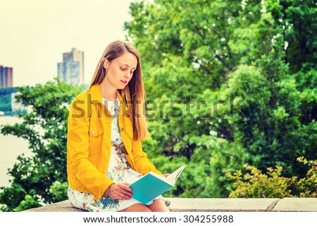 American college student studying in New York. A pretty girl wearing flower patterned underdress, yellow corduroy jacket, holding green book, sitting by trees on campus, looking down, reading.