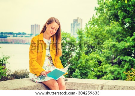 American college student studying in New York. A pretty girl wearing flower patterned underdress, yellow corduroy jacket, holding book, sitting by trees on campus, looking down, smiling, reading.