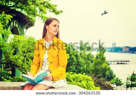 American college student studying in New York, wearing flower patterned underdress, yellow corduroy jacket, holding green book, sitting by Hudson River, reading, thinking. A bird flying on background.