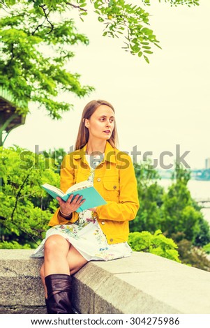 American college student studying in New York. A girl wearing flower patterned underdress, yellow corduroy jacket, holding green book, sitting by trees on campus, reading, thinking. Instagram effect.