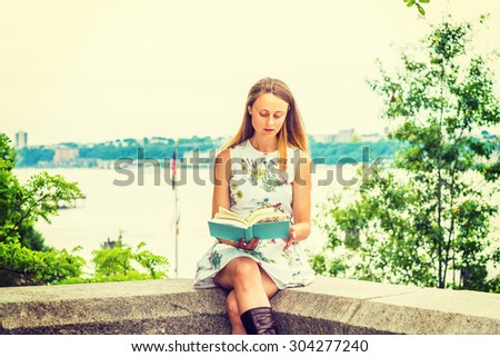 American college student studying, traveling in New York. A pretty girl wearing flower patterned dress, holding green book, sitting by Hudson River, looking down, reading, thinking. Instagram effect.