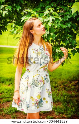 Girl Love Green. Wearing flower patterned white dress, an American college student standing on green lawn by trees on campus, interestedly looking up, thinking. Concept of Environment Protection.