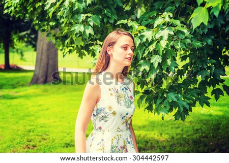 Girl Love Green. Wearing flower patterned white dress, an American college student standing on green lawn by trees on campus, interestedly looking around, thinking. Concept of Environment Protection.