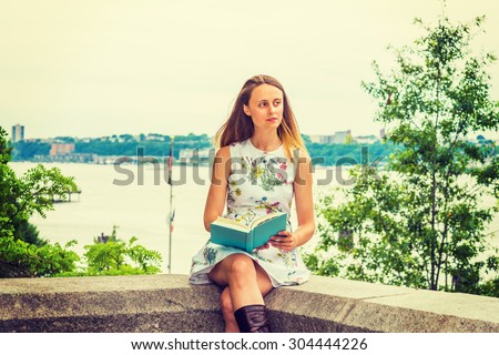American college student studying, traveling in New York. A pretty girl wearing flower patterned dress, holding green book, sitting by Hudson River, looking around, reading, thinking. Instagram effect