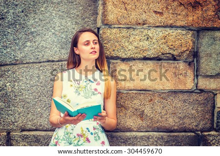 Portrait of American college student. A pretty girl wearing flower patterned, sleeveless, white dress, holding green book, standing by rocky wall, narrowing eyes, reading, thinking. Instagram effect.