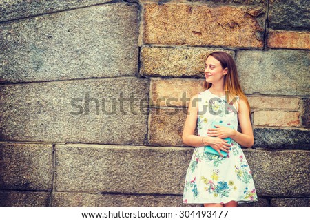 Portrait of College Student. A girl wearing flower patterned, sleeveless, white dress, holding green book, standing against rocky wall on campus, smiling, looking away. Instagram effect. Copy Space.