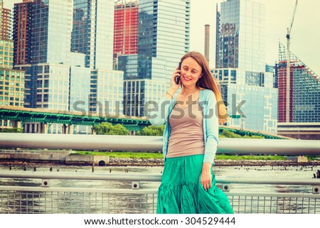 Good to hear from you. Wearing light blue sweater, green skirt, a pretty American woman standing by metal fence on pier in New York, smiling, listening, talking on mobile phone. Instagram effect.