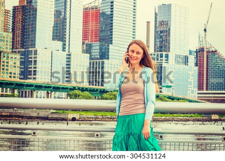Lonely girl wants someone to talk. Wearing light blue sweater, green skirt, an American woman standing by metal fence on pier in New York, sad, depressed, listening to cell phone. Instagram effect.