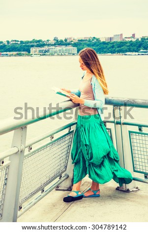American college student wearing light blue cardigan, green skirt, sandals, standing by metal fence by Hudson River in New York, opposite New Jersey, looking down, reading book. Instagram effect.