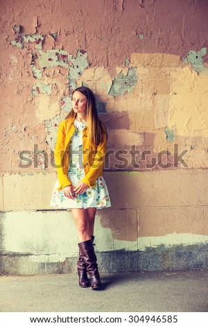 Woman Casual Fashion. A pretty girl wearing flower patterned underdress, yellow corduroy jacket, brown leather high riding boots, standing by wall peeling off paints, looking away. Instagram effect.
