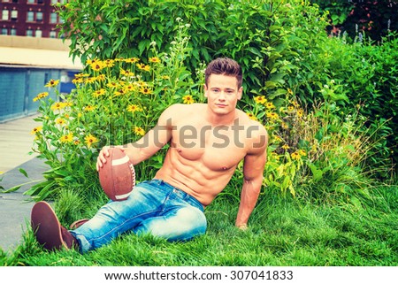 Leisure time in hot summer. Shirtless, half naked, waring jeans, a young sexy guy sitting on green grasses outside in New York, holding American football, smiling, enjoying outdoor activities.