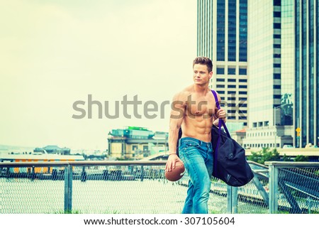 Fitness Model. Man Health. Shirtless, half naked, waring jeans, a young, strong, sexy guy, carrying big duffel bag, holding American football, walking from business district, going to play field.