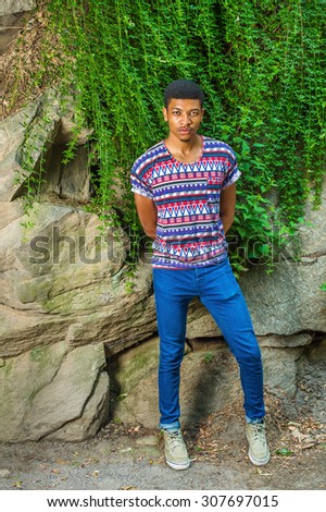 African American Man Casual Fashion in New York. Wearing a colorful pattern shirt, blue jeans, sneakers, a young guy standing by rocks with green leaves, winter jasmine, hands in back, looking at you.