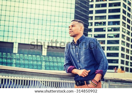 Young African American Man Grown in Big City. Wearing blue shirt, bracelets, a black college student standing in business district with high buildings, hoping, wishing, confidently looking forward.