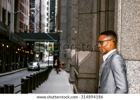 African American Businessman Working in New York. Wearing gray blazer, glasses, a black college student standing on street, sad, thinking, lost in thought. A car driving on narrow road on background.