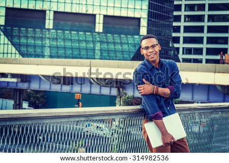African American college student studying in New York. Wearing blue shirt, glasses, bracelets, holding laptop computer, a young black man standing in business district with high buildings, smiling.