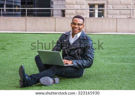 African American college student studying in New York. Wearing leather jacket, jeans, leather shoes, glasses, a young black man sitting on green lawn on campus, working on laptop computer, smiling.