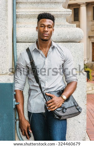 Man Casual Urban Fashion. Wearing gray shirt, jeans, wristwatch, carrying shoulder leather bag, an African American guy standing in the corner of street, listening music with earphone and cell phone.