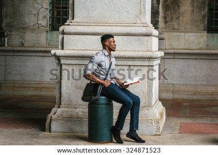 African American College Student studying in New York. Wearing gray shirt, jeans, cloth shoes, carrying shoulder leather bag, a black man sitting on metal pillar on street, reading book, thinking.