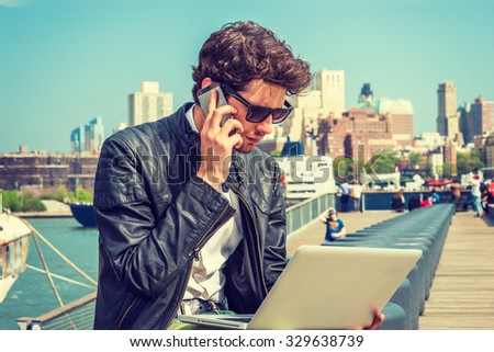 Businessman traveling, working in New York. Wearing sunglasses, a young guy with beard, sitting on bench at harbor, working on laptop computer, talking on phone in the same time. Instagram effect.
