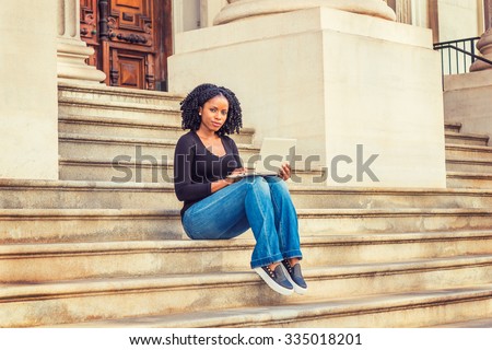 African American college student studying in New York. Wearing long sleeves, V neck top, jeans, leather sneakers, a black girl sitting on stairs outside office on campus, working on laptop computer.