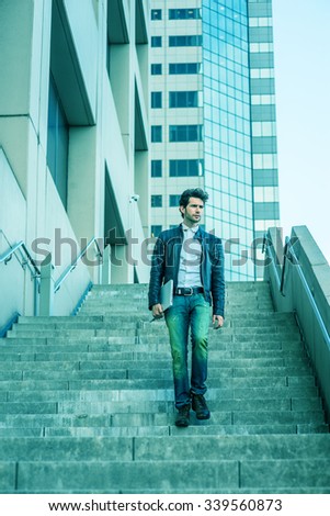 Young man with beard, wearing leather jacket, jeans, arm carrying laptop computer, walking down stairs outside office high building after busy working day, going home. Filtered look with cyan tint.